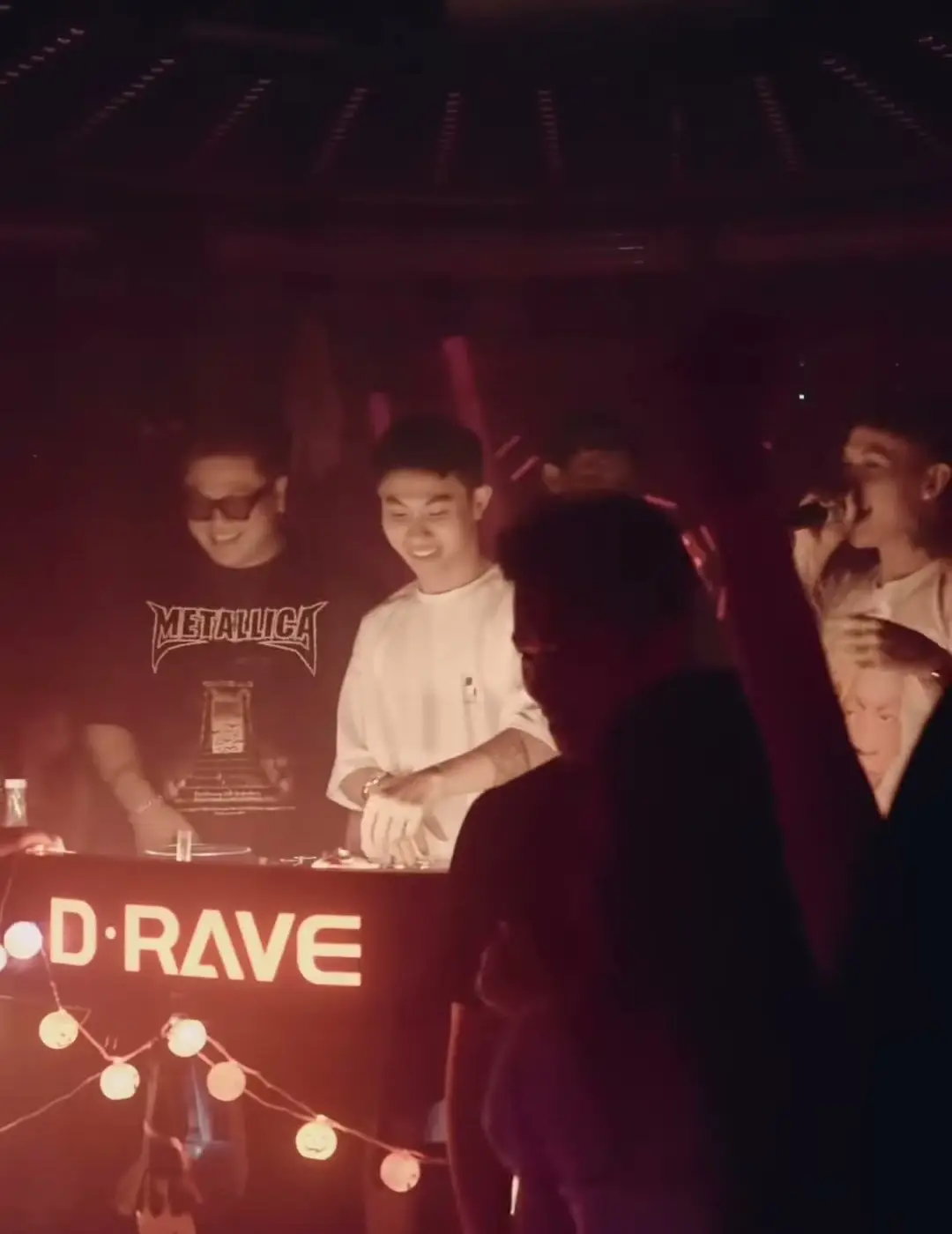 Hai DJ sau bốt có chữ D·RAVE phát sáng, dây đèn tròn trang trí phía trước